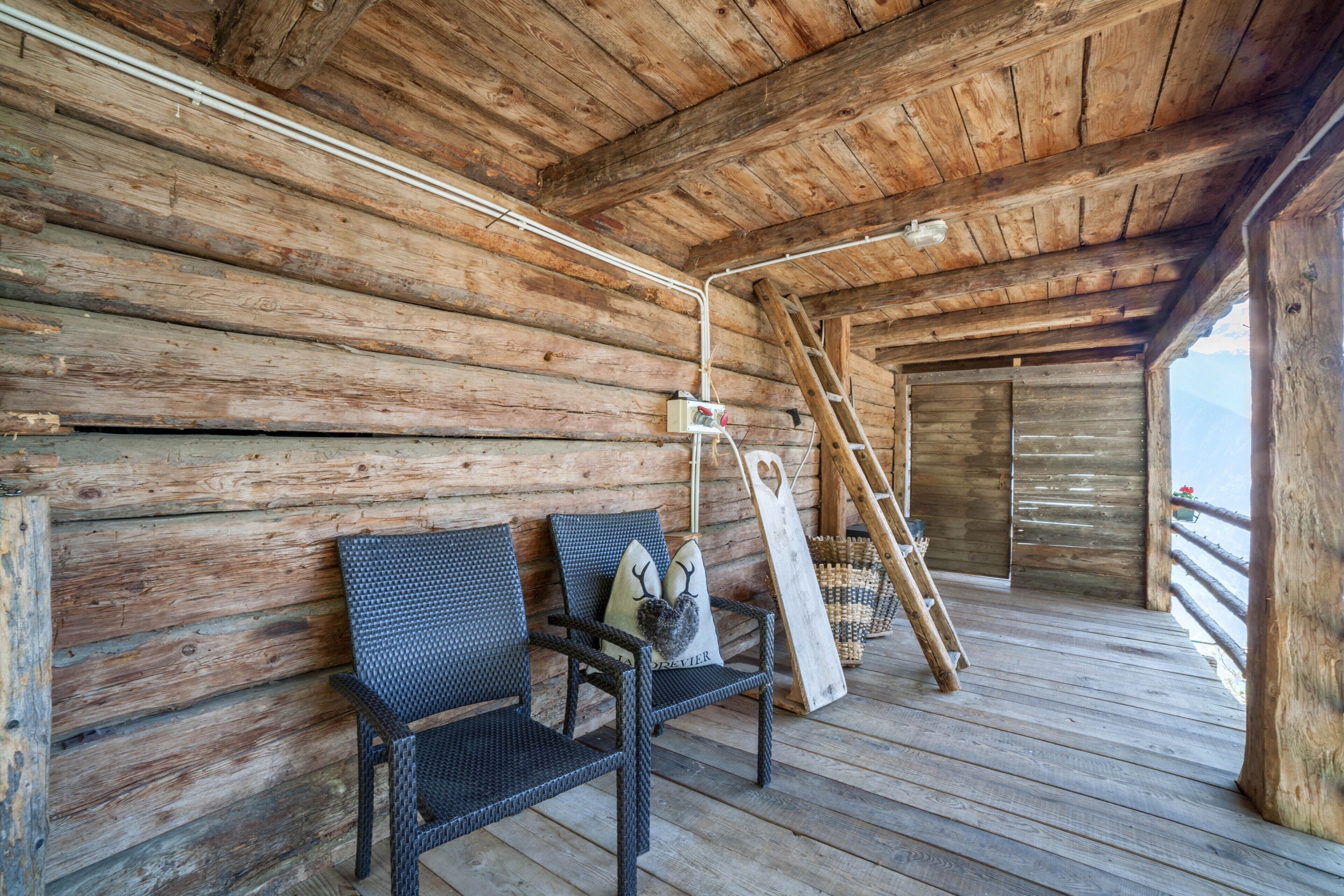A wooden cabin interior with a wooden floor, a bench, a ladder, and a window overlooking a mountainous landscape.