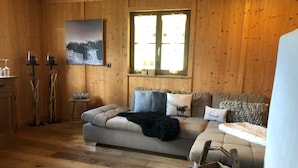 L’intérieur chaleureux d’un chalet avec un plancher en bois, un canapé agrémenté de plusieurs coussins, une fenêtre avec des stores et un tableau accroché au mur.