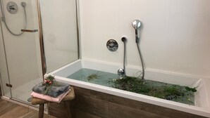 Une salle de bain moderne avec une douche à parois de verre, un tabouret en bois sur lequel sont posées des serviettes et une baignoire remplie de plantes.