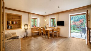 A room with a wooden floor, a dining table with chairs, a bookshelf, a flat-screen TV, and a large window with a view of trees. - Apartment “Schildhofsuite” in “Schildhof Ebion” with Mountain View & Garden (St. Leonhard Passeier)