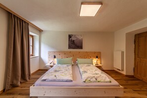 A double bed with a headboard, two bedside tables with lamps, a window with curtains, a radiator, and a wooden door. - Apartment “Familienwohnung” in “Schildhof Ebion” with Mountain View, Wi-Fi & Garden (St. Leonhard Passeier)