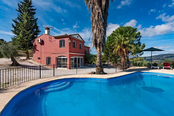 A red villa with a swimming pool, surrounded by trees and a clear sky.