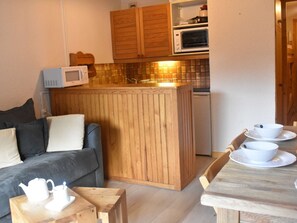 A compact living space with a kitchenette, a wooden dining table with white cups and saucers, a grey sofa with cushions, and a small wooden side table with a teapot and cup.