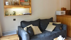 A living room with a dark grey sofa, wooden shelves with various items, a white radiator, and a microwave on a wooden counter. - Ski-In/Ski-Out Studio for 4 in Méribel, Pool Access, Near Shops & Bus Stop (Méribel)
