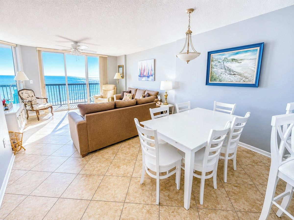 A spacious living area with a white dining table and chairs, a beige sofa, and a balcony with a view of the ocean.