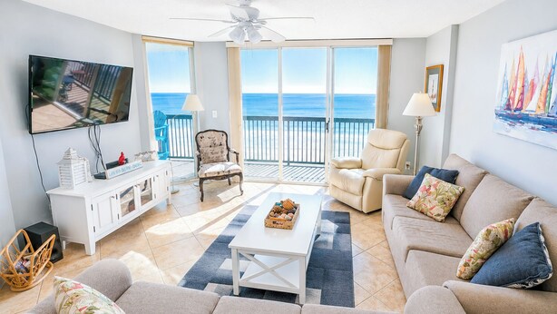 Een woonkamer met een bank, fauteuil, salontafel, televisie en een balkon met uitzicht op zee.