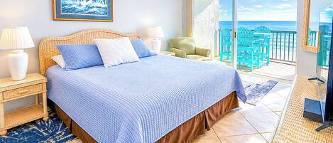 Un dormitorio con cama, mesitas de noche, ventilador de techo, un ventanal con vistas al océano y una silla de mimbre.