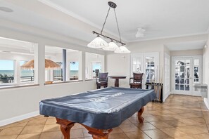 Una mesa de billar en una habitación con ventanas amplias, un ventilador de techo y vistas al océano.