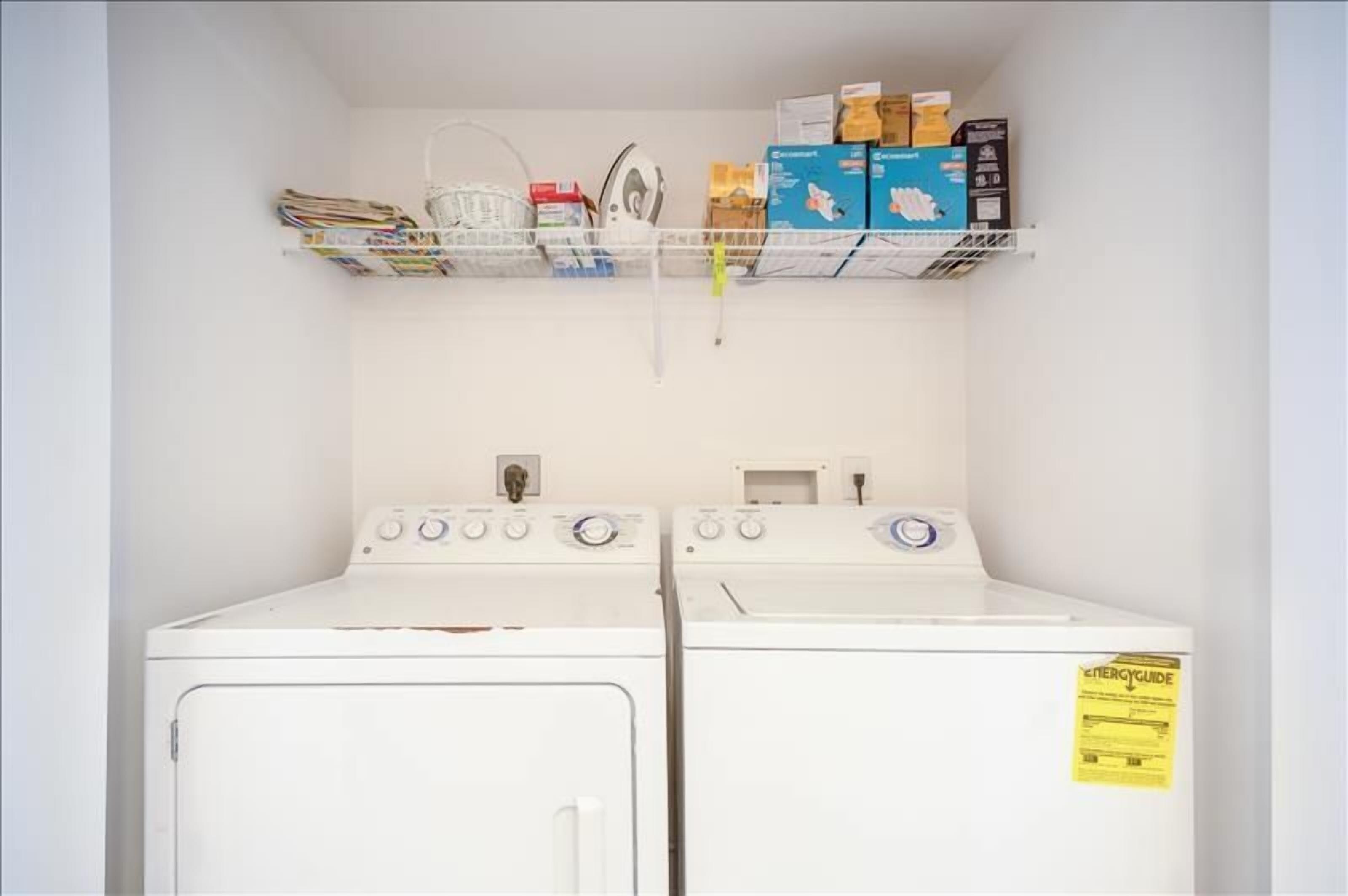 Une buanderie avec deux machines à laver blanches et une étagère au-dessus, sur laquelle sont rangés divers objets.