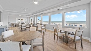 Un comedor amplio con varias mesas y sillas, ventanas grandes que ofrecen vistas al océano y ventiladores de techo.