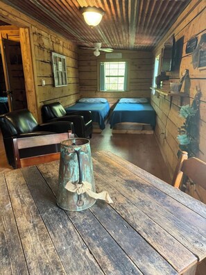A rustic cabin interior with a wooden table, a metal pitcher, two beds, and a television.