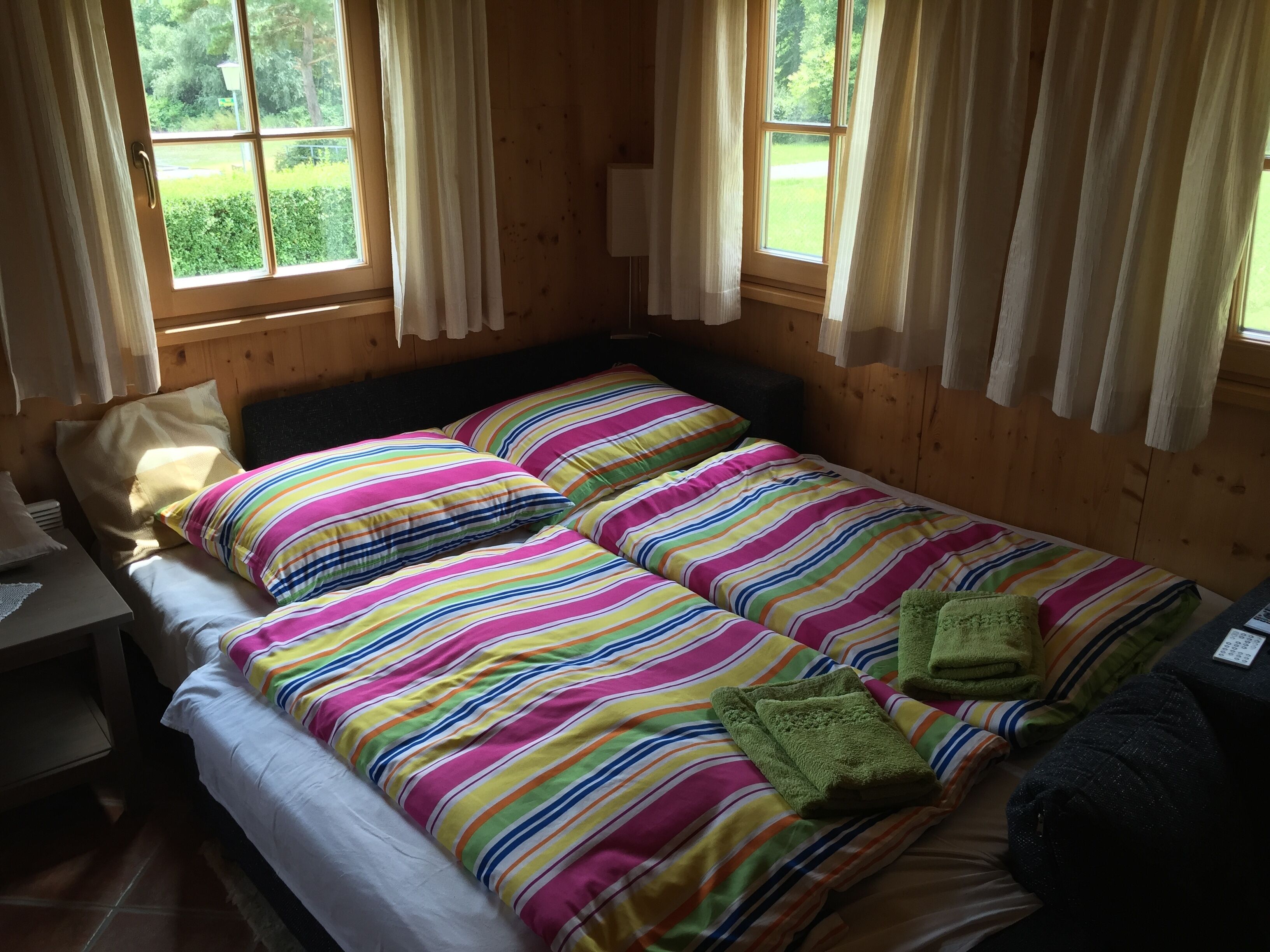 A room with two beds, each with colorful striped bedding, a small table, and a window with curtains.