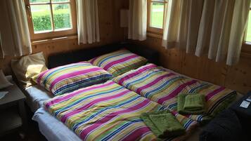 A room with two beds, each with colorful striped bedding, a small table, and a window with curtains.