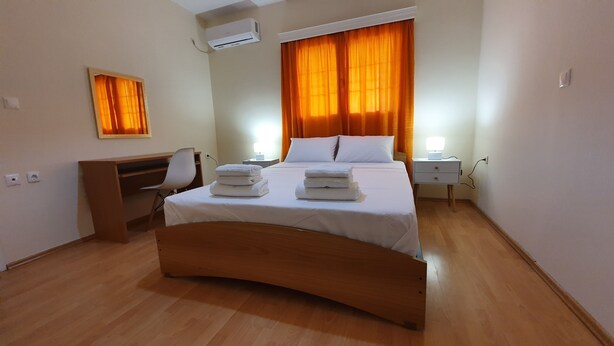 A hotel room with a bed, a desk, a chair, an air conditioner, and a nightstand with a lamp. - MTC-SK Apartment by the sea and Airport  (Markopoulo Mesogaias)