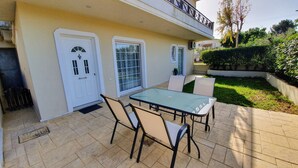 A patio with a glass table and chairs, a white door, and a small garden area. - MTC-SK Apartment by the sea and Airport  (Markopoulo Mesogaias)
