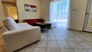 A living room with a white sofa, a red sofa, a black coffee table, and a white ottoman. There is a painting on the wall and a window with a geometric pattern. - MTC-SK Apartment by the sea and Airport  (Markopoulo Mesogaias)