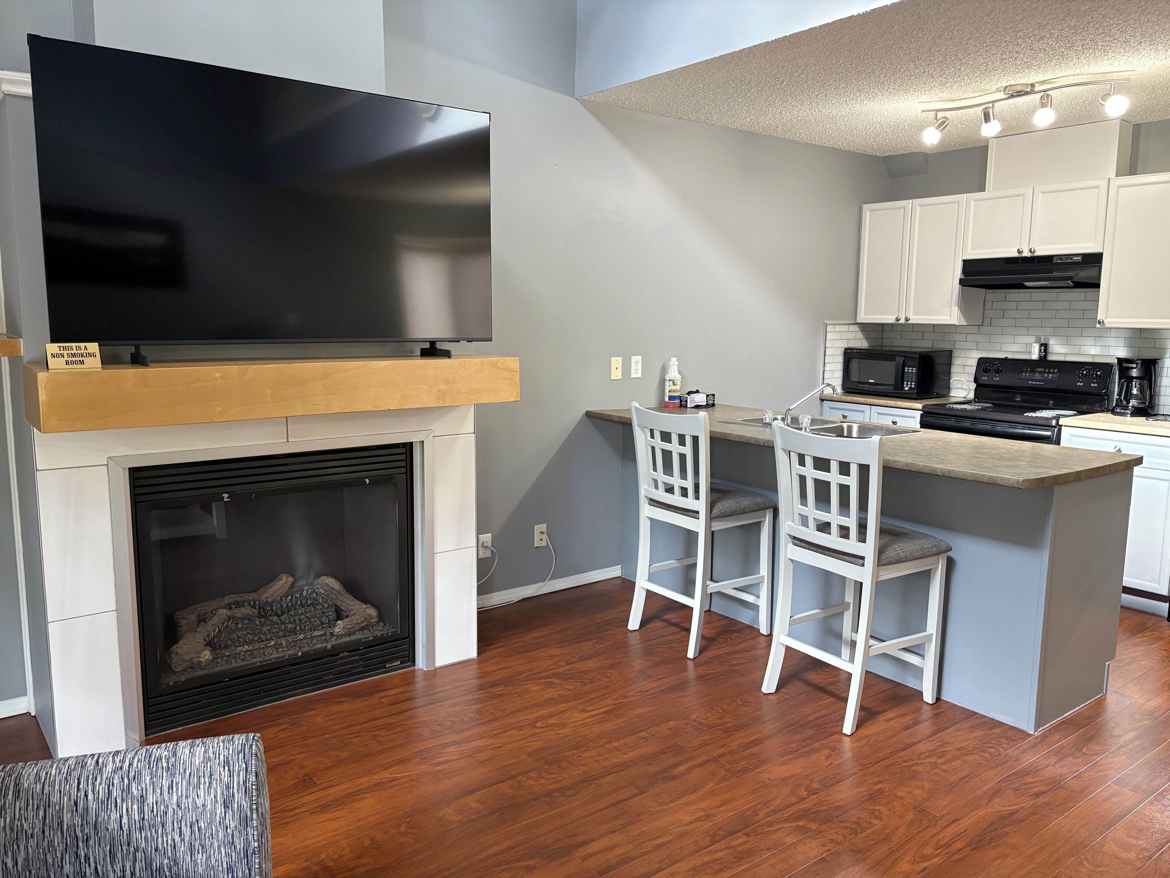 A modern kitchen with a flat-screen TV, a fireplace, white bar stools, and a built-in island with a sink.