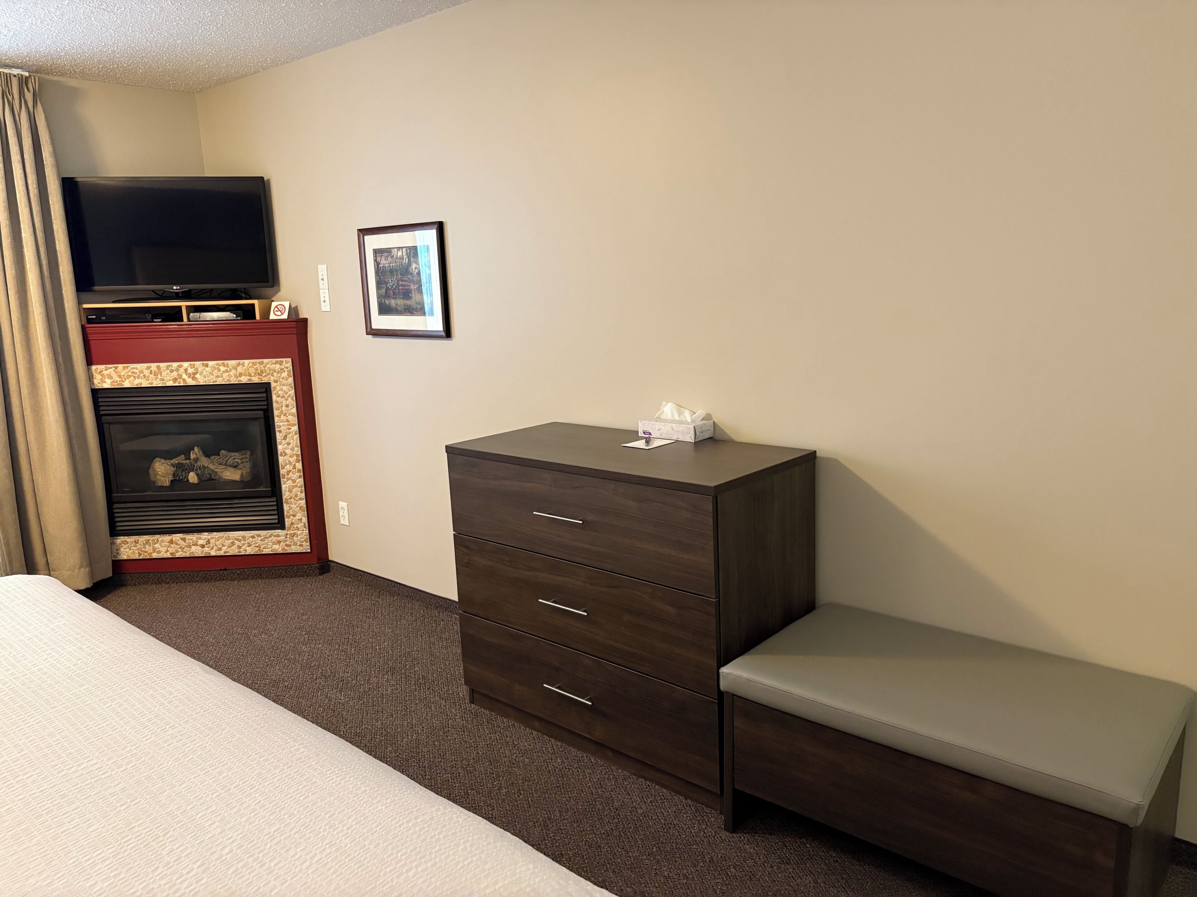 Una habitación de hotel con cama, cómoda, banco, chimenea y televisión.