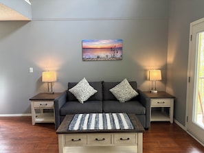 A living room with a gray sofa, two patterned pillows, a woven coffee table, and a sunset painting on the wall. - Village Creek Country Inn (Westerose)