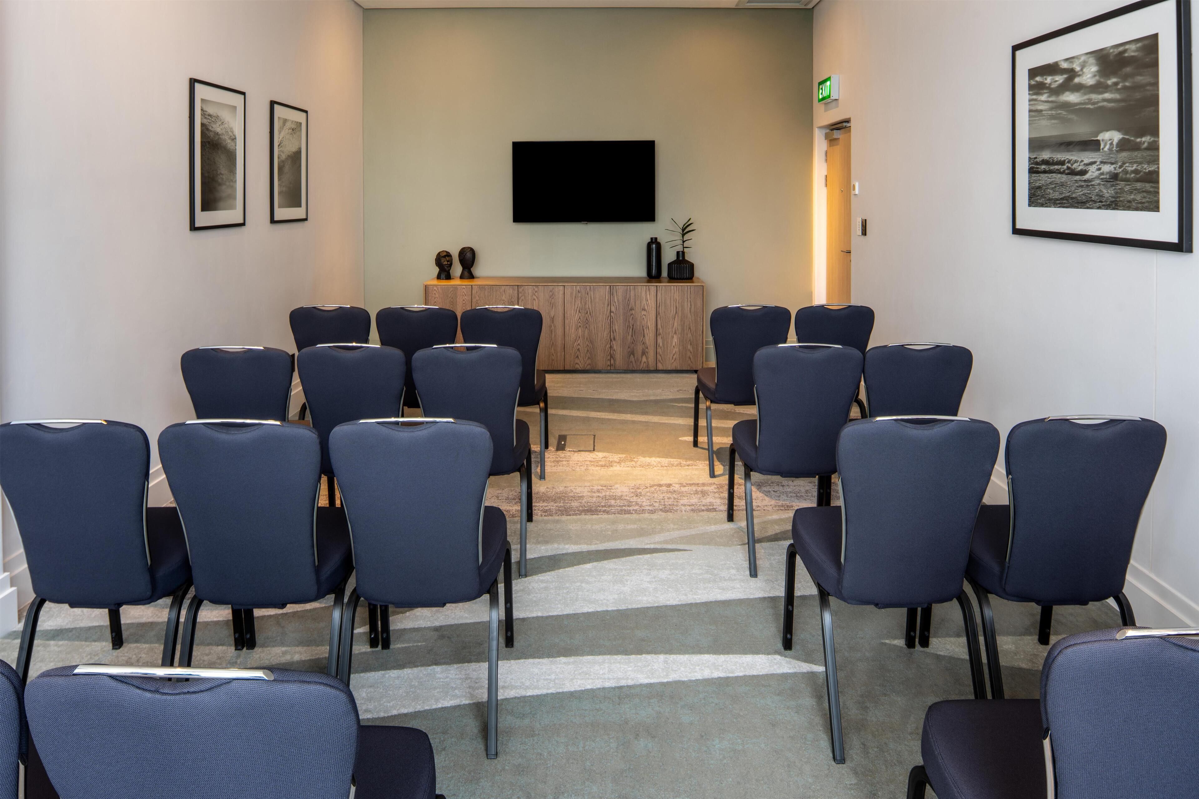 Een vergaderzaal met rijen stoelen, een flatscreen televisie, omlijste kunstwerken en een houten kast.