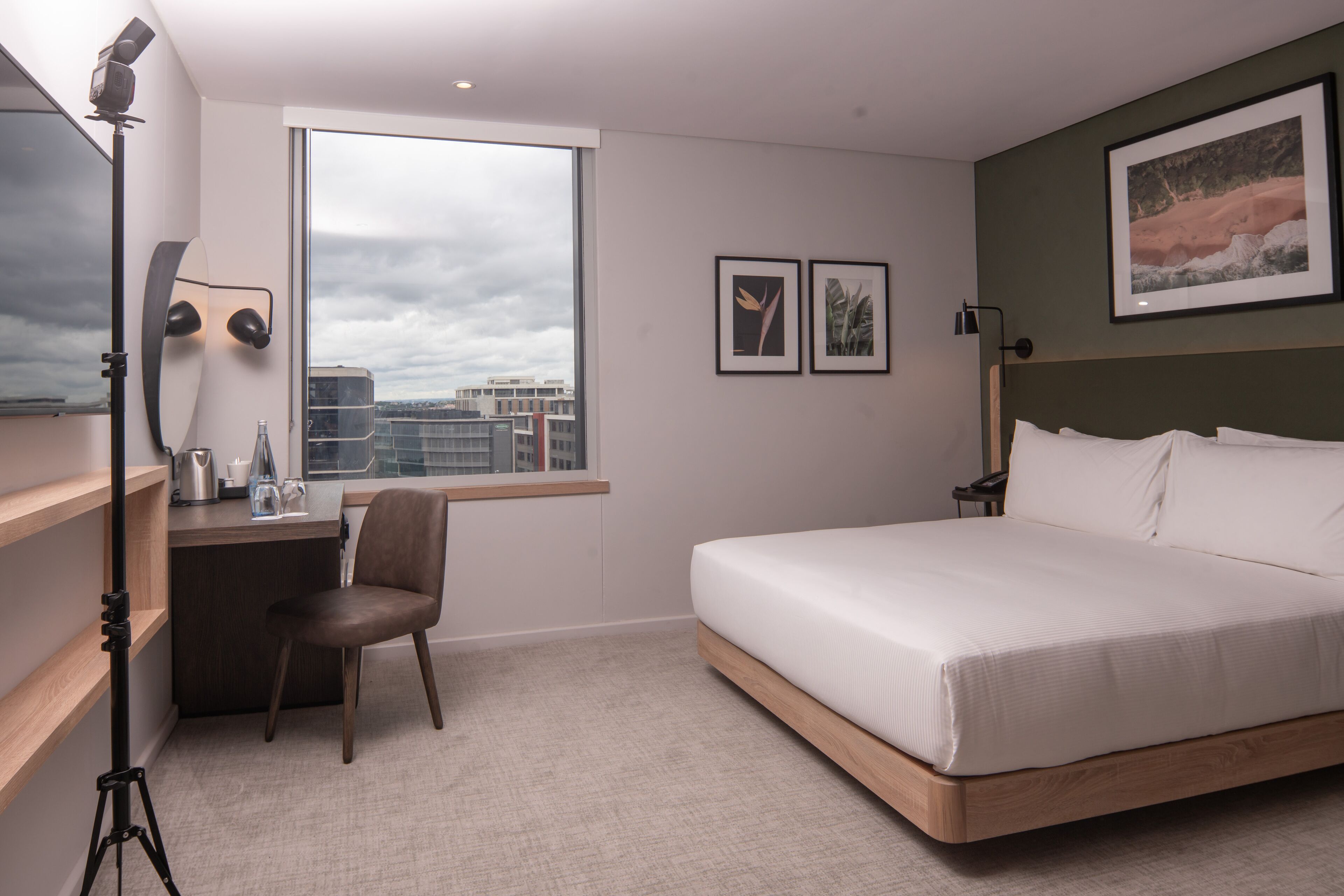 A modern hotel room with a large bed, a desk with a chair, a window with a city view, and a mounted camera.