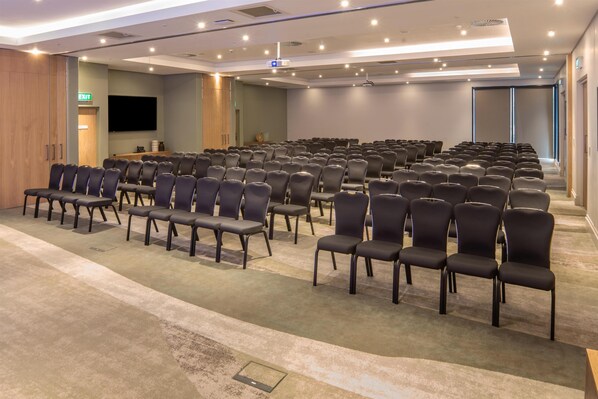Un salón de conferencias con hileras de sillas, una pantalla de proyector y un televisor de pantalla plana.