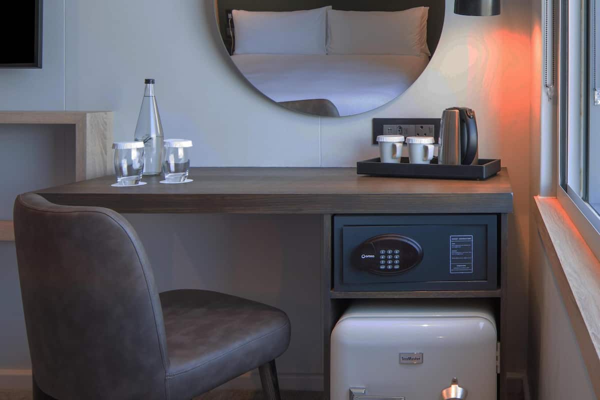 A compact hotel room with a bed, a desk with a chair, a small refrigerator, a bedside table with a lamp, and a large mirror.