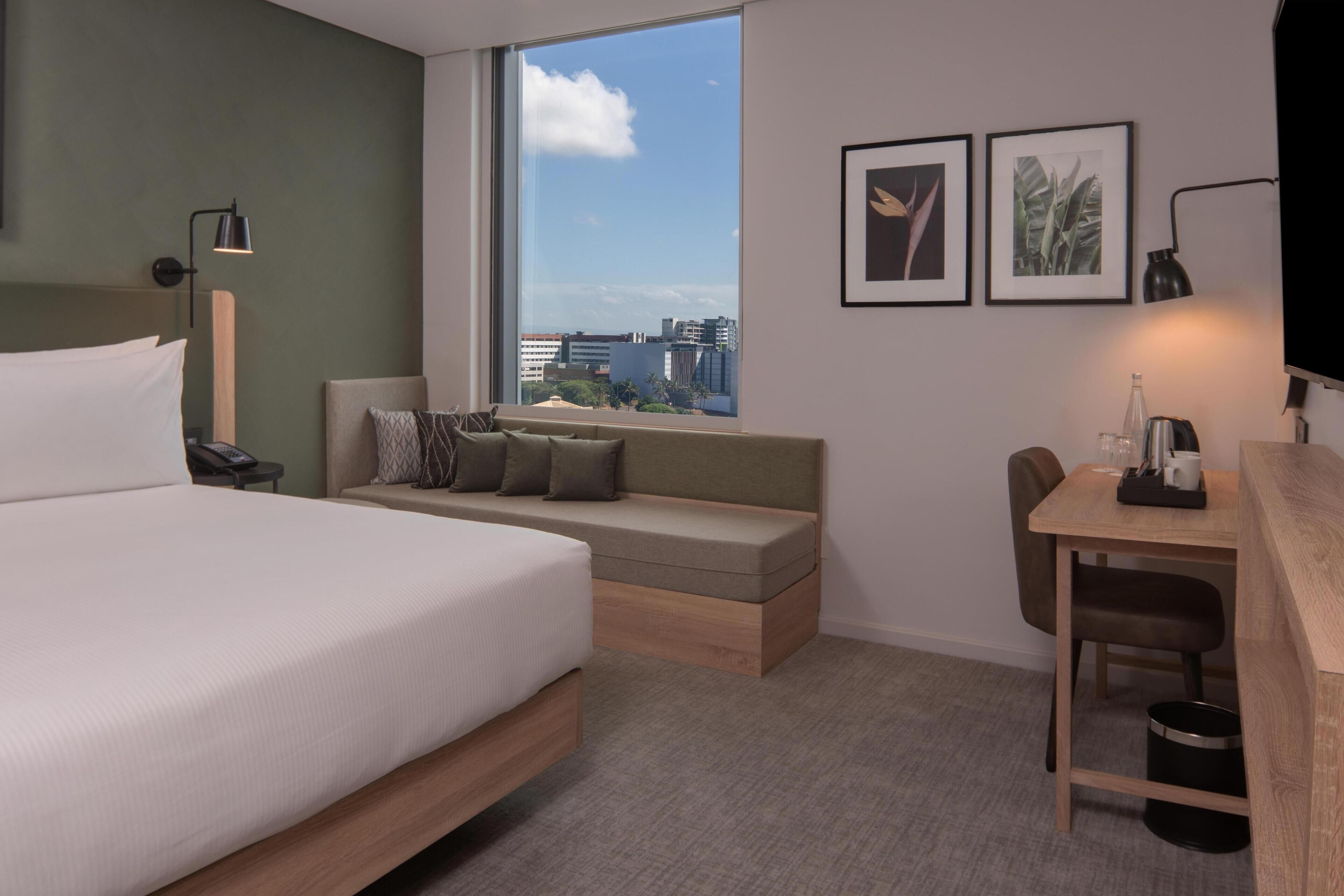 A modern hotel room with a bed, a sofa bed, a desk, and a large window offering a city view.