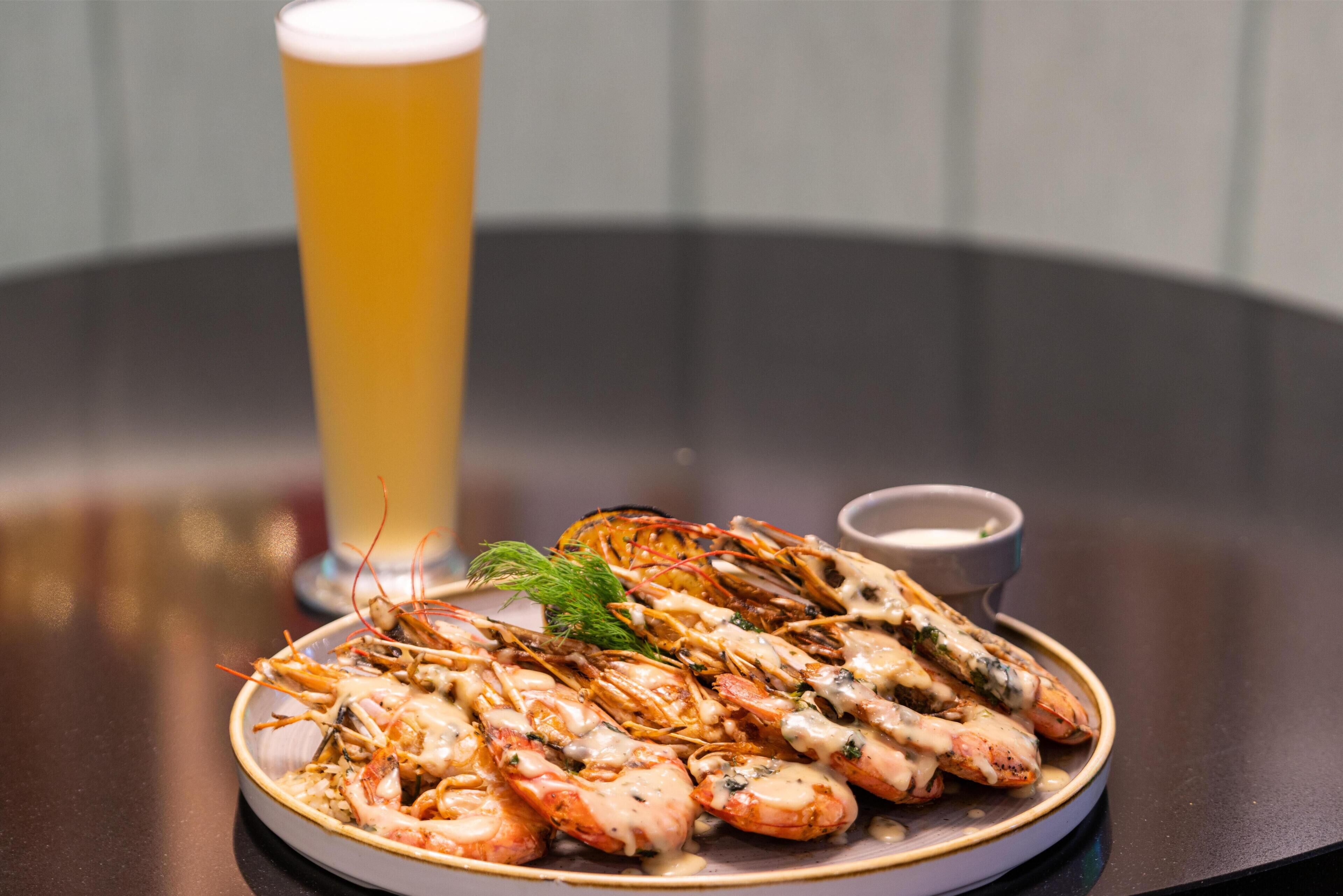 Ein Teller mit gegartem Fisch und Meeresfrüchten, ein Glas Bier und eine kleine Schale Soße auf einem runden Tisch.
