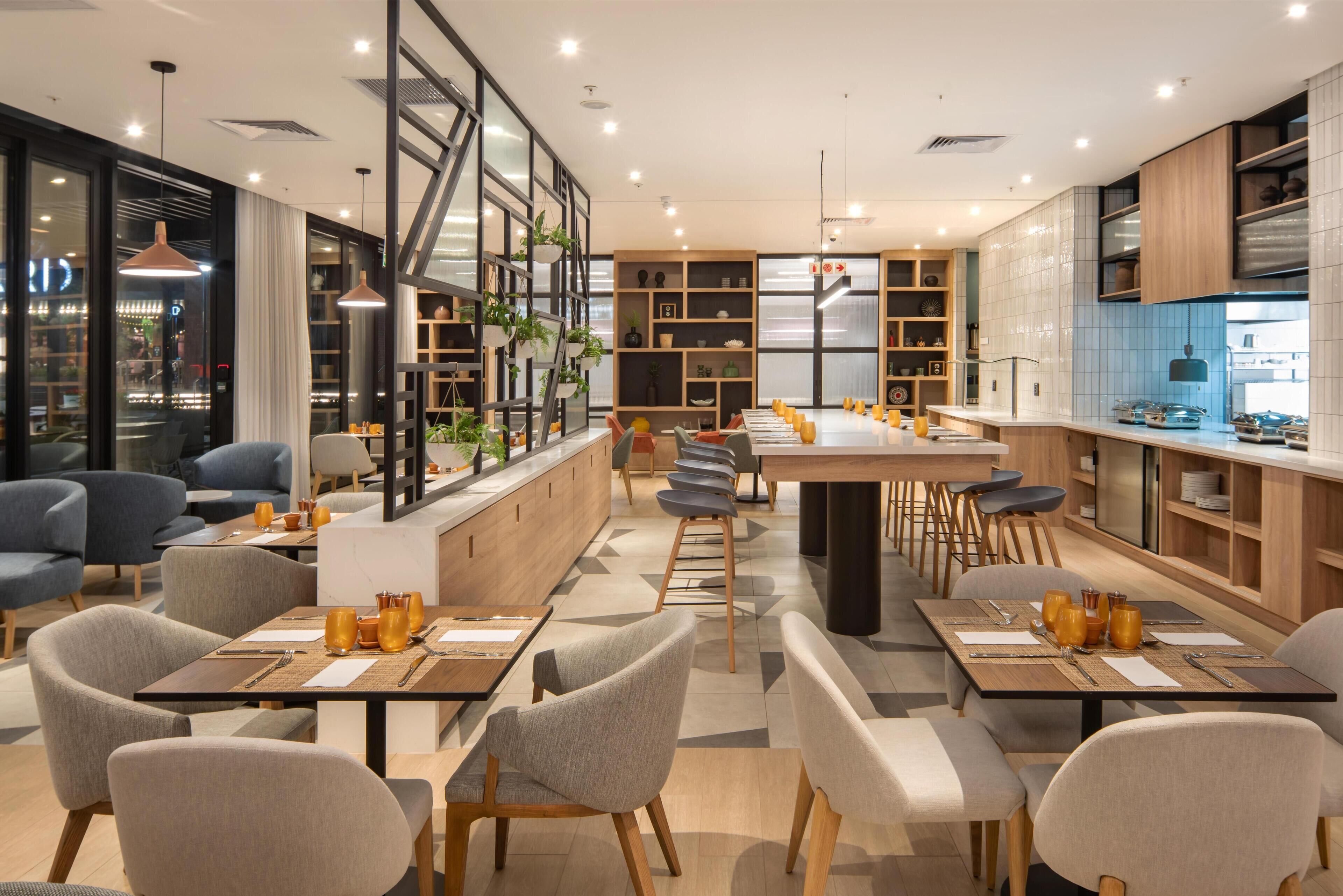 A modern restaurant with a long communal dining table, individual seating, and a kitchen area visible through glass partitions.