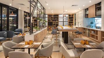 A modern restaurant with a long communal dining table, individual seating, and a kitchen area visible through glass partitions.