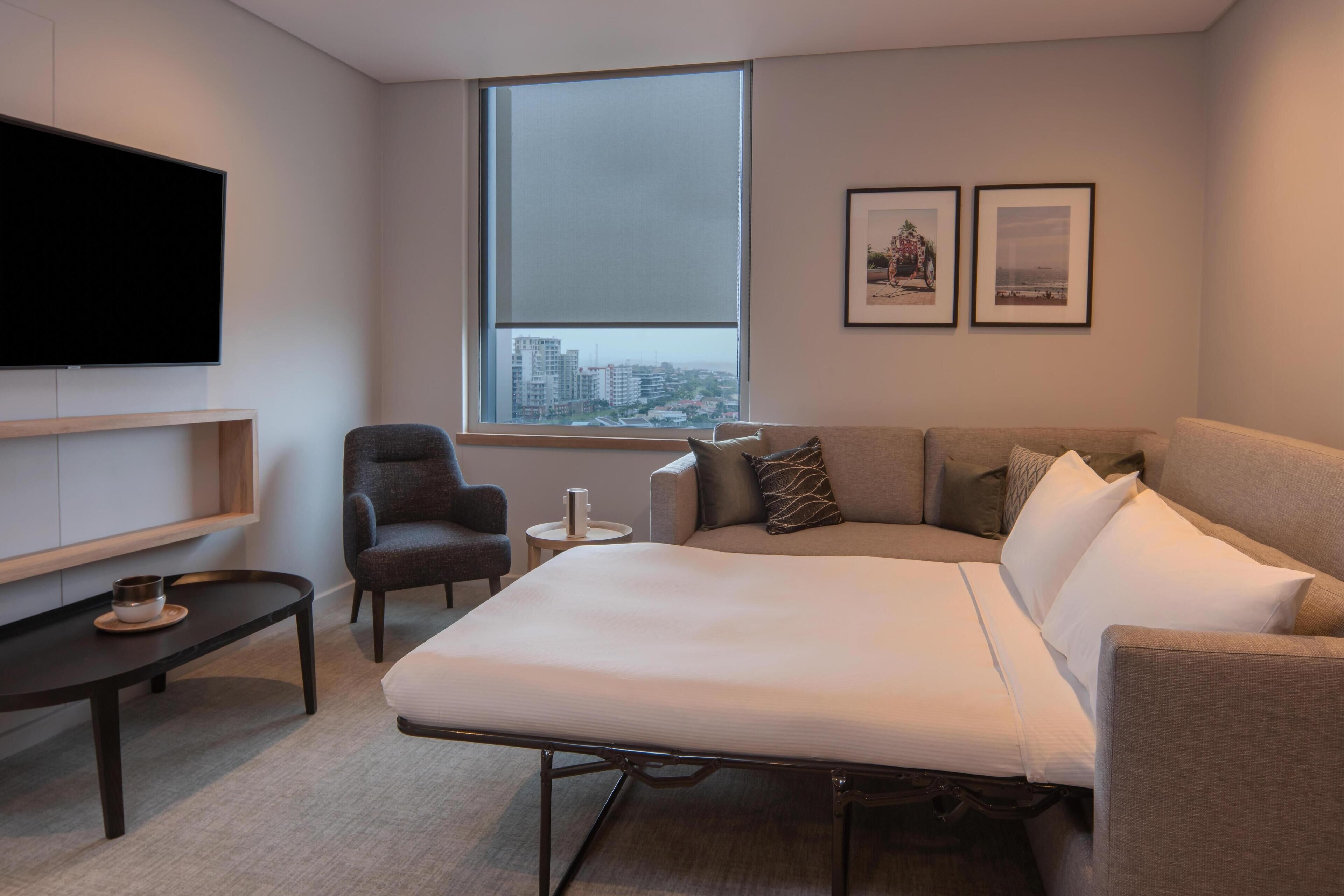 A modern hotel room with a bed, a TV, a small table, a chair, and a window with a city view.