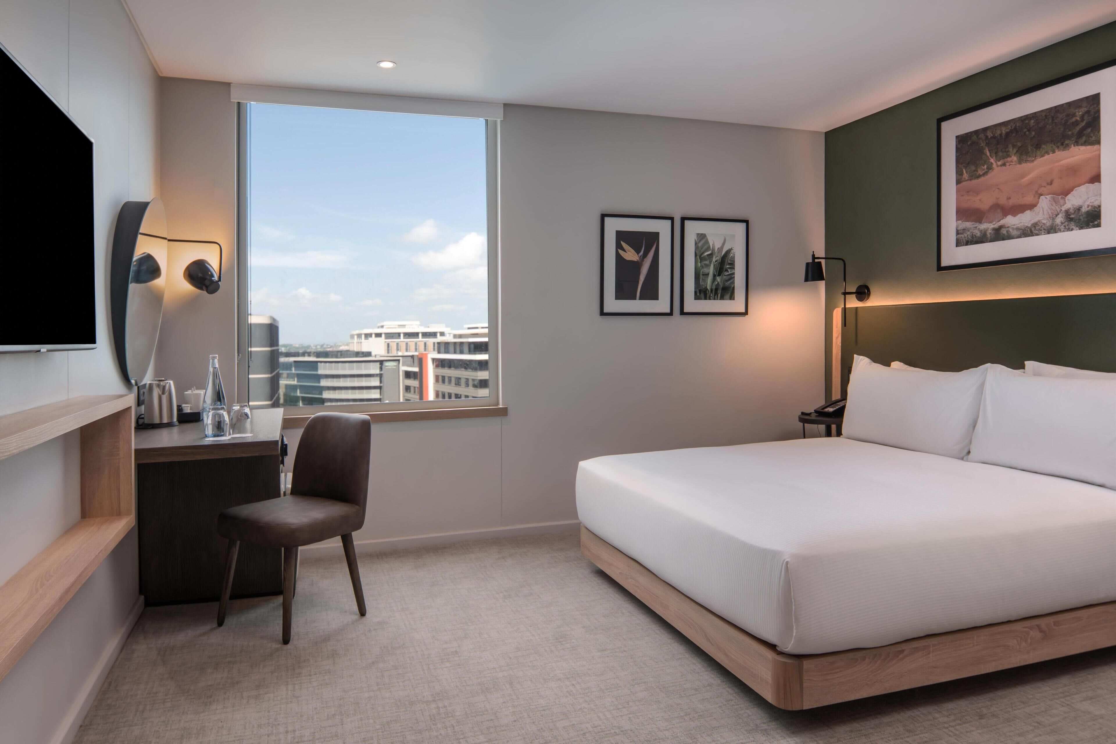 Habitación de hotel con cama, escritorio, silla y un amplio ventanal con vistas a la ciudad.