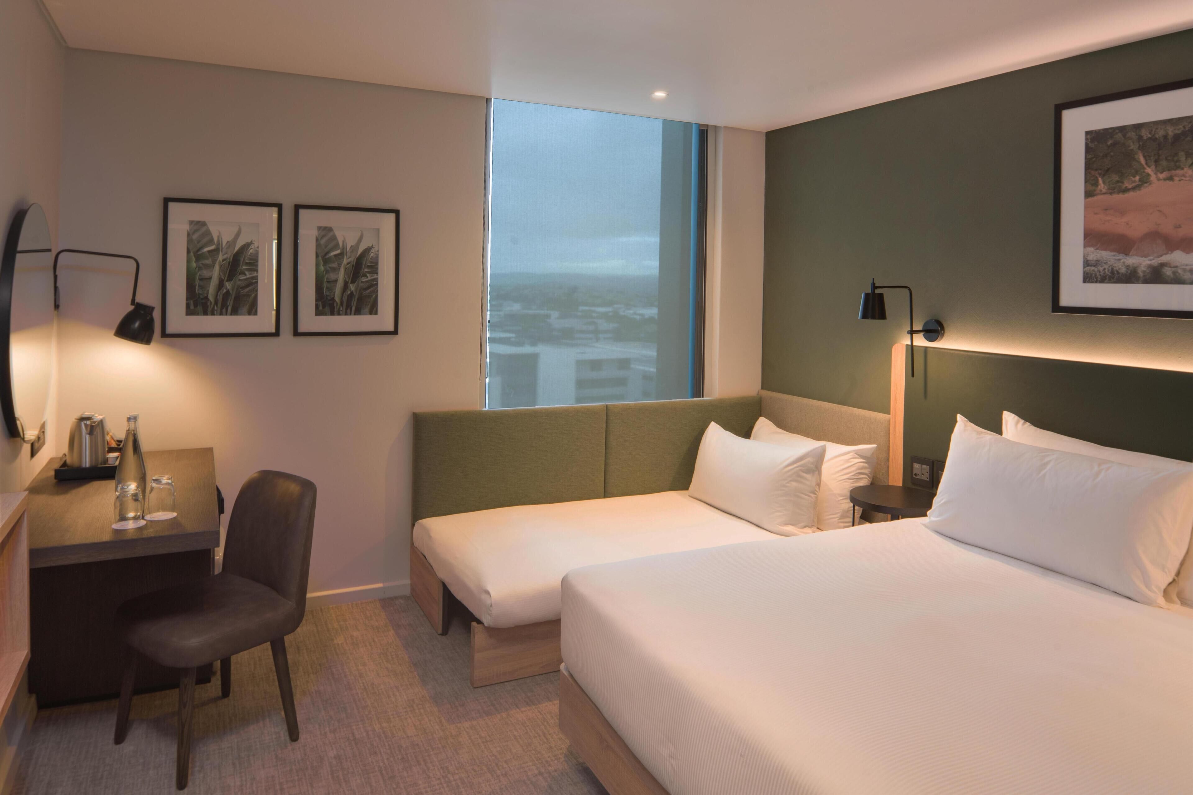 Une chambre d’hôtel avec un lit, un bureau, une chaise, une grande fenêtre et deux tableaux encadrés accrochés au mur.