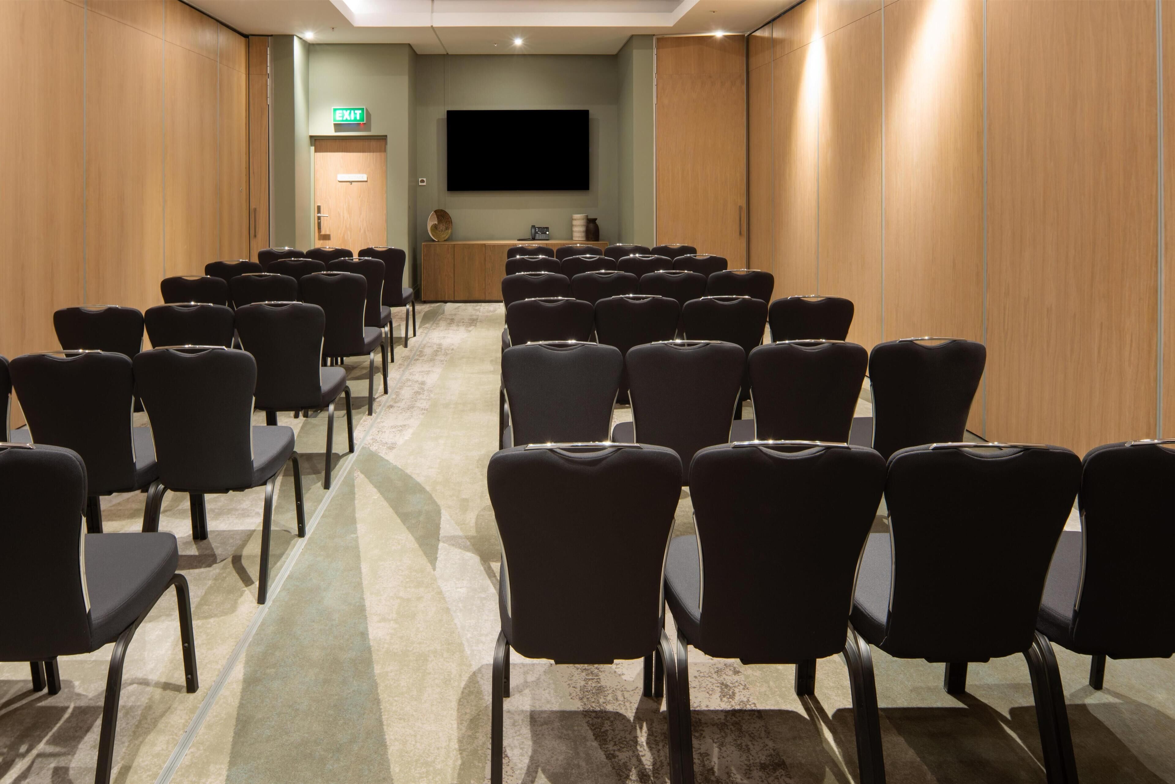 A conference room with rows of chairs facing a flat-screen TV, wooden panel walls, and a carpeted floor.