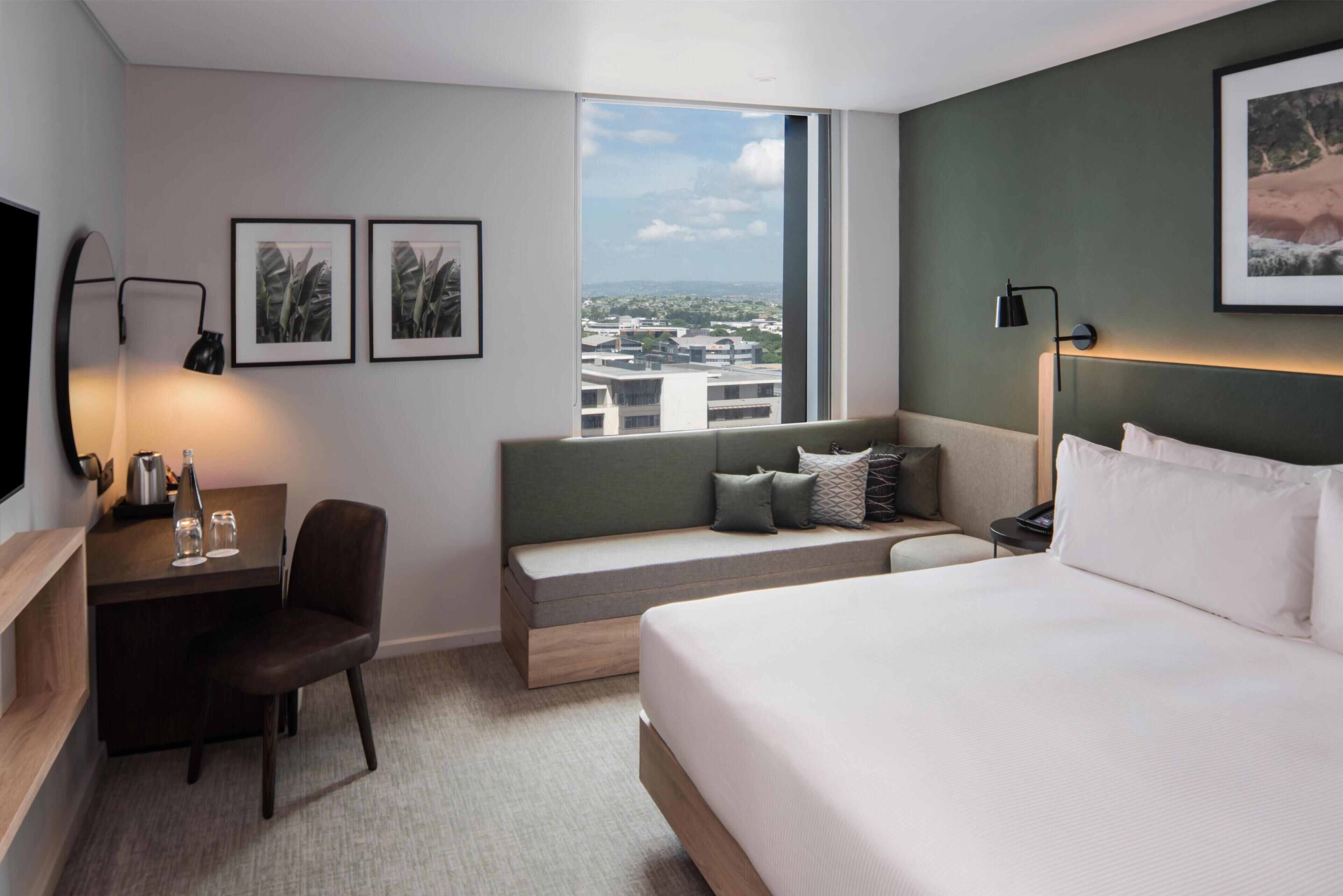 A hotel room with a bed, a desk with a chair, a bench, a TV, and framed pictures on the wall.