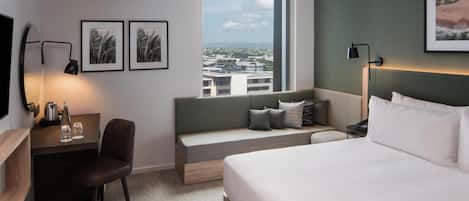 A hotel room with a bed, a desk with a chair, a bench, a TV, and framed pictures on the wall.