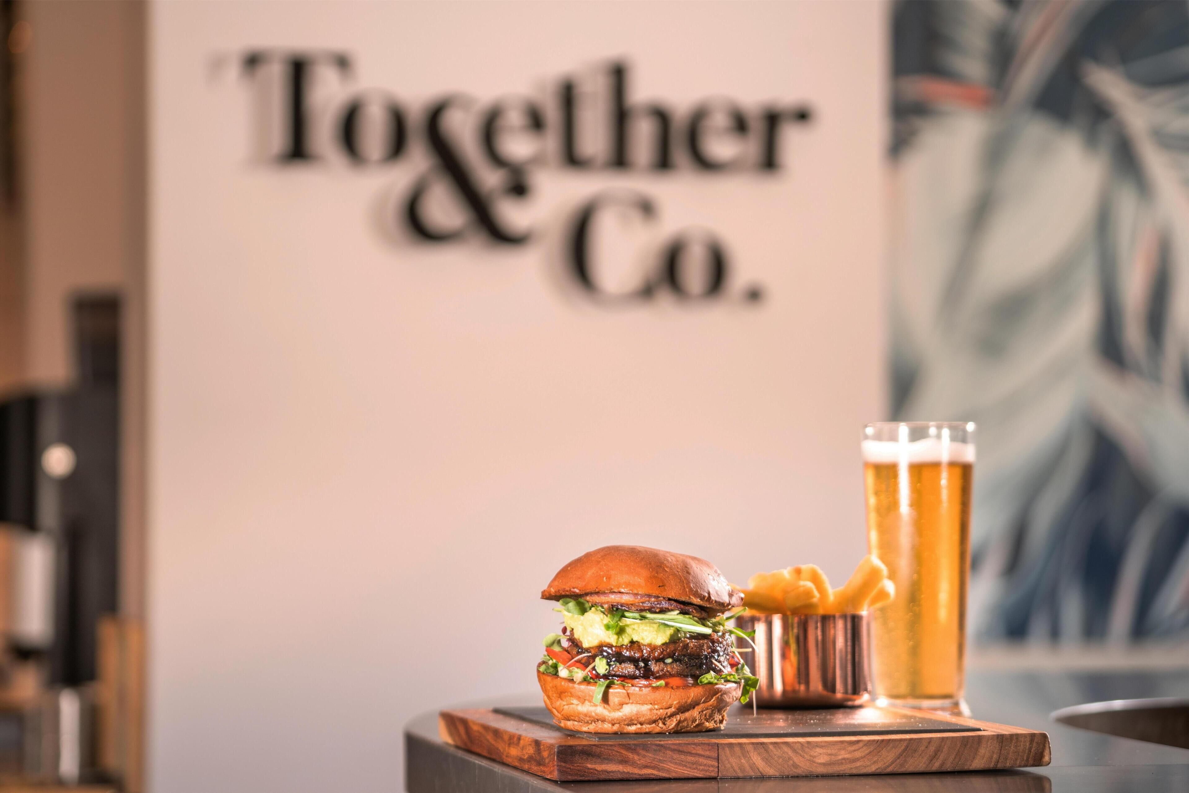 Ein Burger, ein Bierglas und Pommes auf einer Holzplatte, im Hintergrund ein Schild.