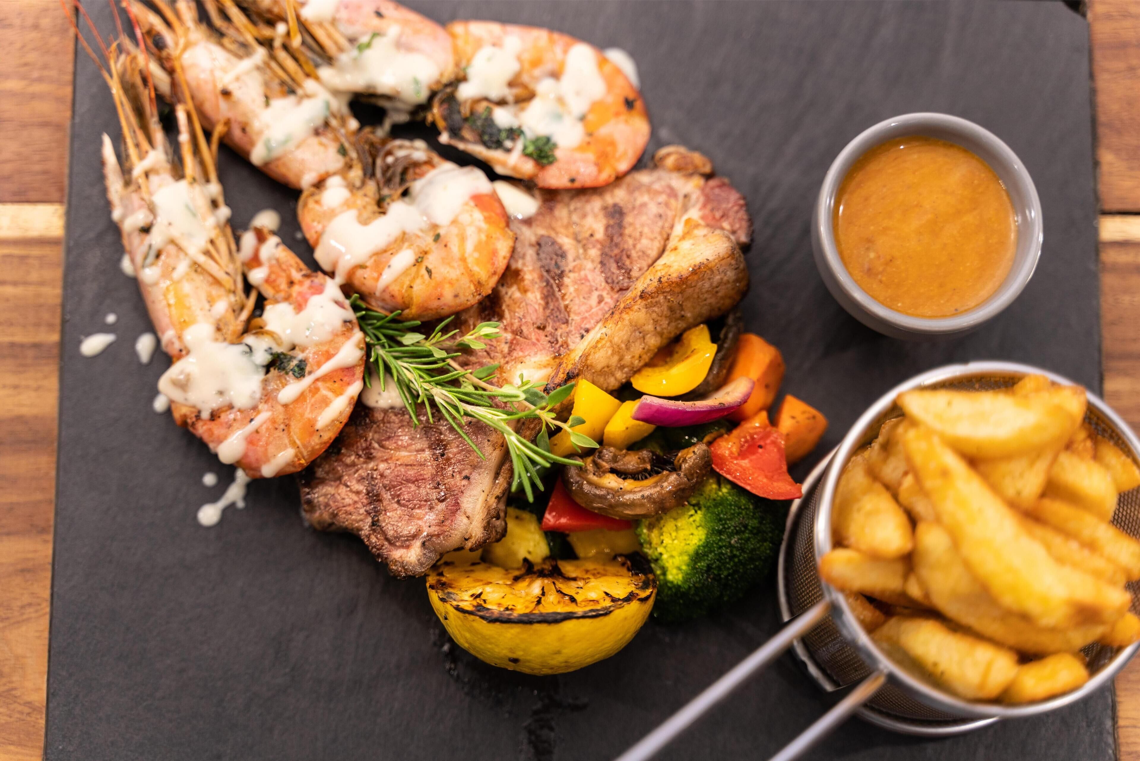 A meal with grilled meat, shrimp, vegetables, and fries on a slate board.