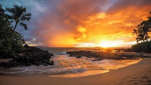 Uma praia com afloramentos rochosos e palmeiras ao pôr do sol.