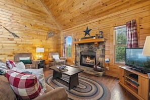 L’intérieur d’une cabane en rondins, avec une cheminée en pierre, des murs en bois, un téléviseur et un coin salon confortable.