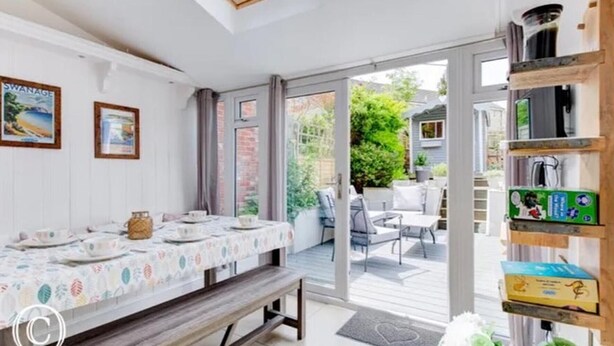 A dining area with a table set for four, a bench, and a view of the garden through glass doors. - Charming 3-bed Cottage in Swanage (Swanage)