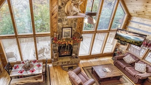 A spacious living room with a stone fireplace, wooden furniture, and large windows offering a forest view. - Campfire Lodge is a beautiful cabin with all the special touches needed to make your stay memorable! (Sevierville)