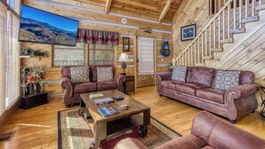 A log cabin interior with a living room featuring two leather sofas, a coffee table, and a television displaying a mountain view. - Campfire Lodge is a beautiful cabin with all the special touches needed to make your stay memorable! (Sevierville)