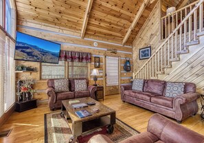 A log cabin interior with a living room featuring two leather sofas, a coffee table, and a television displaying a mountain view.