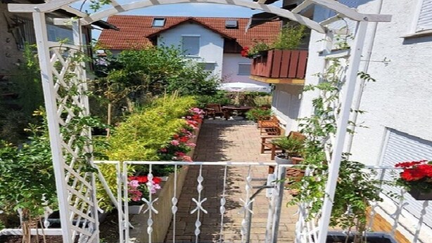 Un sendero de jardín que lleva a una puerta blanca con un banco, rodeado de plantas y flores en macetas.