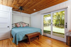 Ein Schlafzimmer mit Holzboden, einem Bett mit türkisgrünlicher Bettdecke, einer Bank und einer Glastür zum Garten.