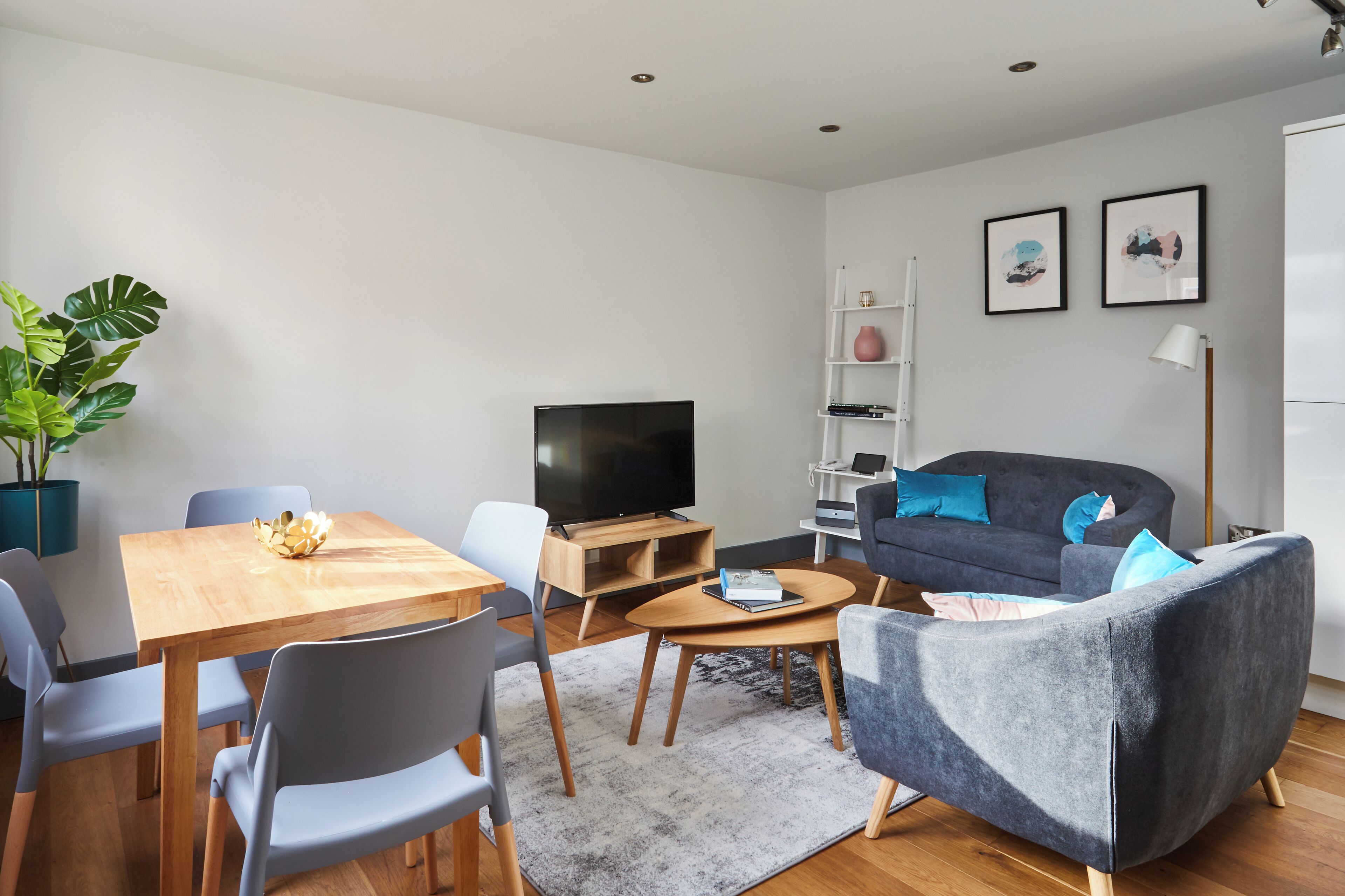 A modern living room with a wooden dining table, grey chairs, a grey sofa, a flat-screen TV, and a potted plant.