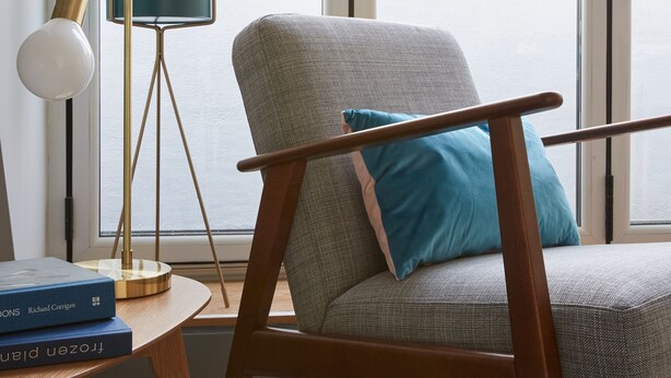 A modern living room with a grey armchair, a teal pillow, a floor lamp, and a potted plant by a large window.