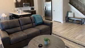 Un salon moderne avec un canapé en cuir marron, une table basse agrémentée d’une plante, et un coin cuisine doté de meubles en bois foncé et d’appareils électroménagers en acier inoxydable.